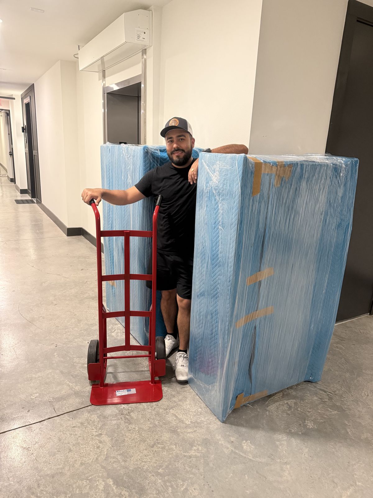 An American Outlaw Logistics mover walking a shrink-wrapped piece of furniture out on a hand truck during a Fort Worth move