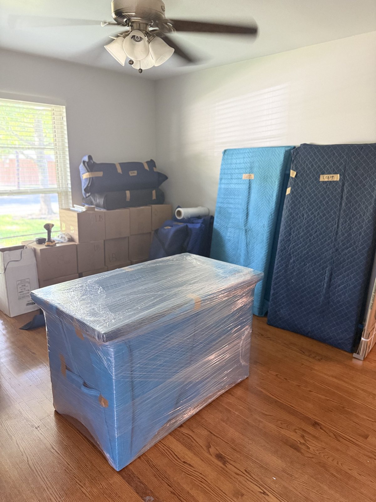 A Fort Worth home mid-pack: labeled cartons stacked, a dresser shrink-wrapped over moving blankets, and padded pieces staged against the wall